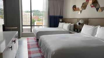 Quarto de hotel com duas camas, uma mesa, uma cadeira, janela com vista para edifícios e uma obra de arte na parede.