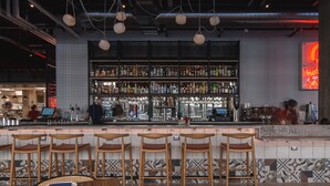 Una zona de bar con varios taburetes, una barra repleta de botellas y un letrero de neón.