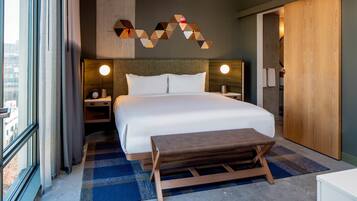 Quarto de hotel moderno com uma cama grande, mesas de cabeceira, um banco de madeira, um detalhe decorativo na parede com formas geométricas e vista pela janela.