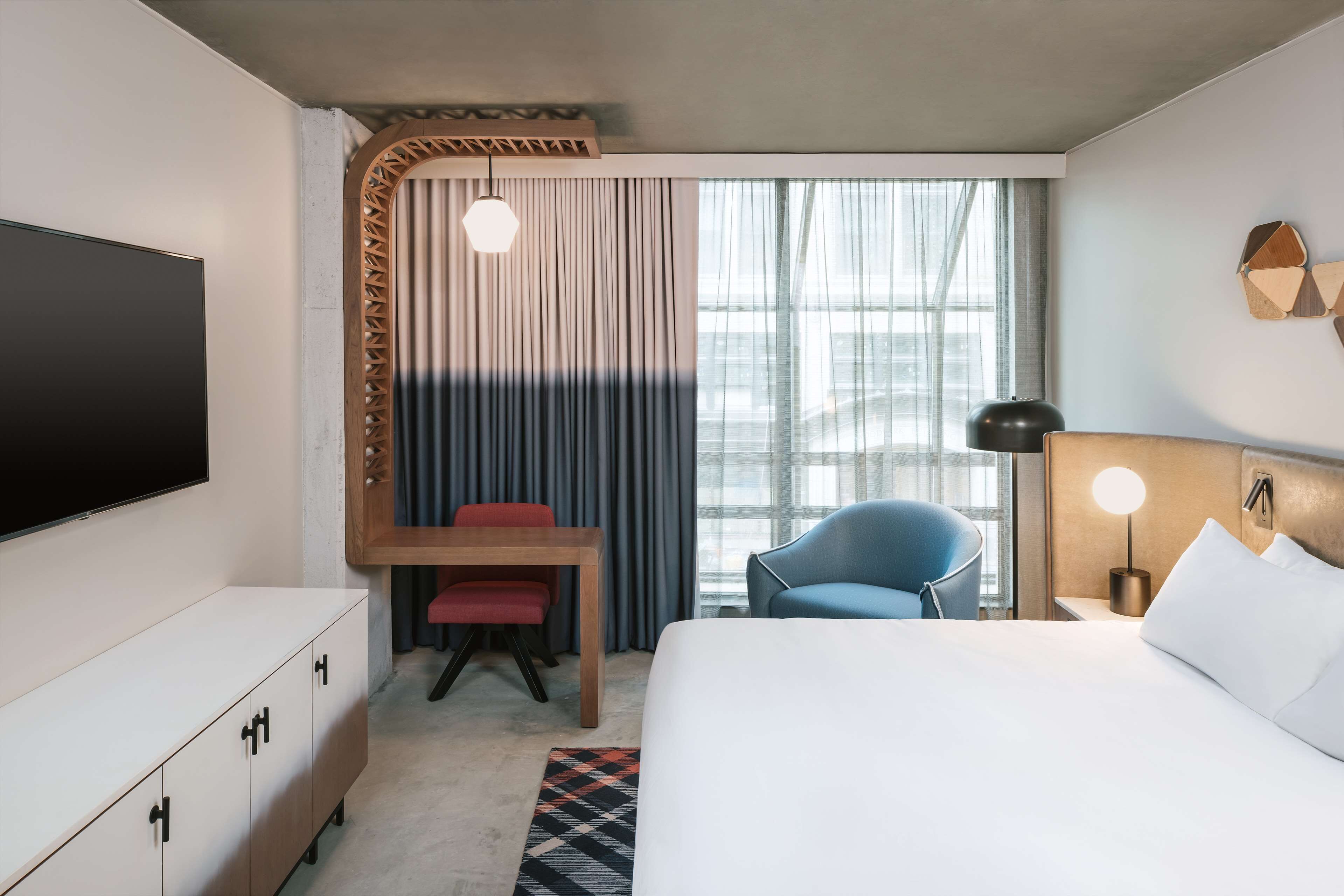 Une chambre d’hôtel avec un lit, une télévision, un bureau avec une chaise, un fauteuil bleu et une fenêtre avec des rideaux.