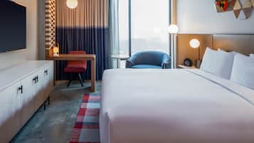A hotel room with a large bed, a desk, a chair, a TV, and a view of the city.