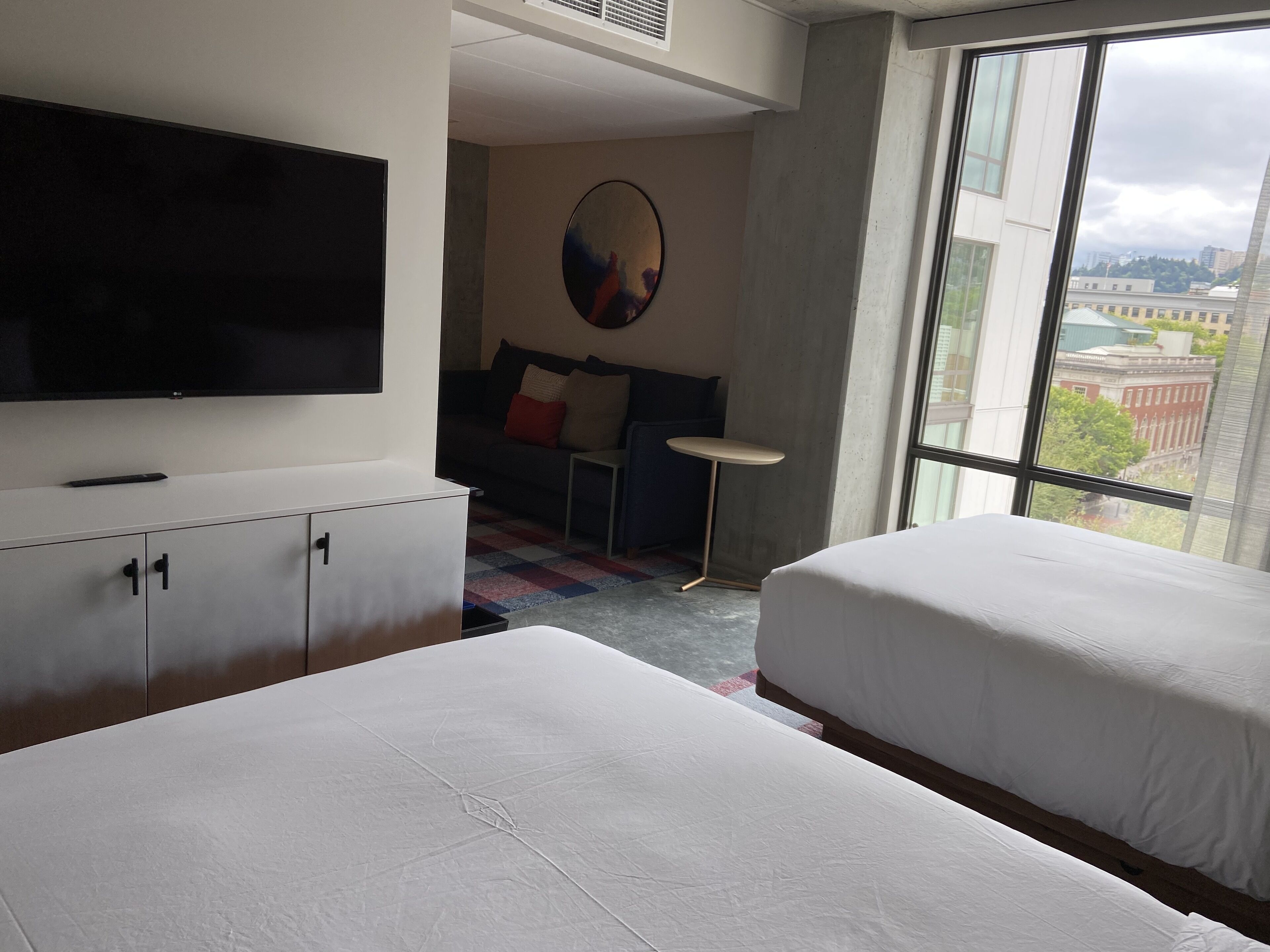 Ein Hotelzimmer mit Bett, Fernseher, Sofa, kleinem Tisch und großem Fenster mit Blick auf die Stadt.
