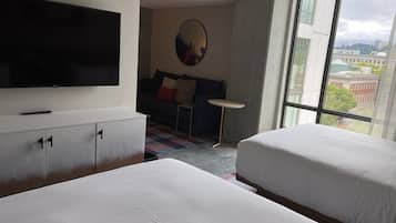 A hotel room with a bed, a TV, a sofa, a small table, and a large window offering a city view.