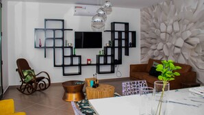 Una sala de estar moderna con un televisor de pantalla plana, estanterías negras, un sofá marrón, un sillón amarillo y una pared decorativa con un diseño geométrico.