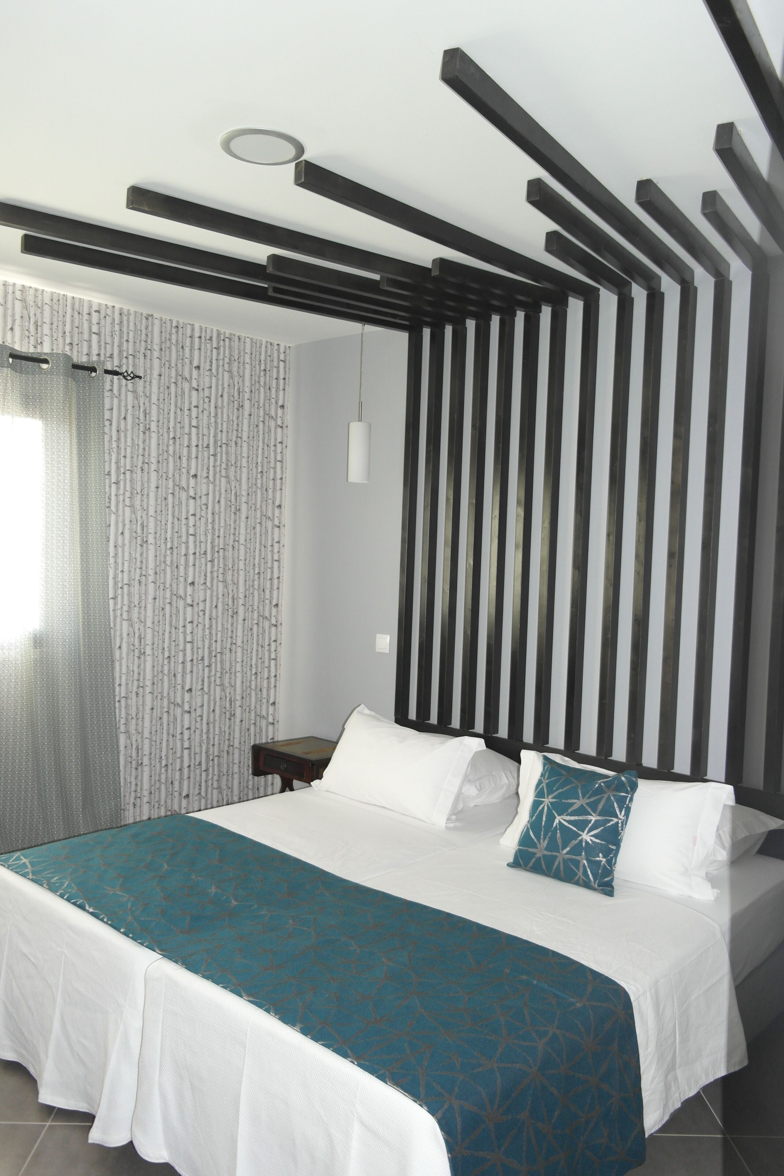 A modern bedroom with a bed, white bedding, a teal-patterned throw, and a vertical black and white striped wall.