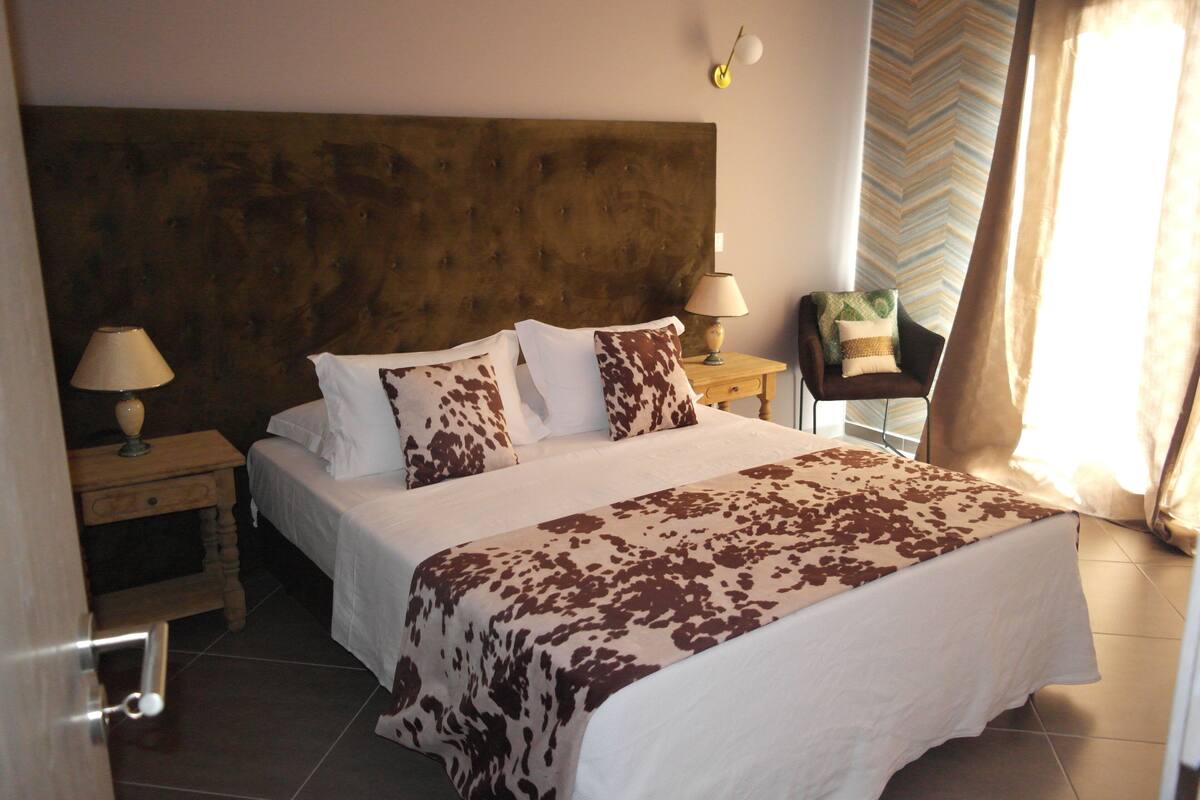 A neatly made bed with a patterned bedspread, two bedside tables with lamps, a chair with a cushion, and a chevron-patterned wall.