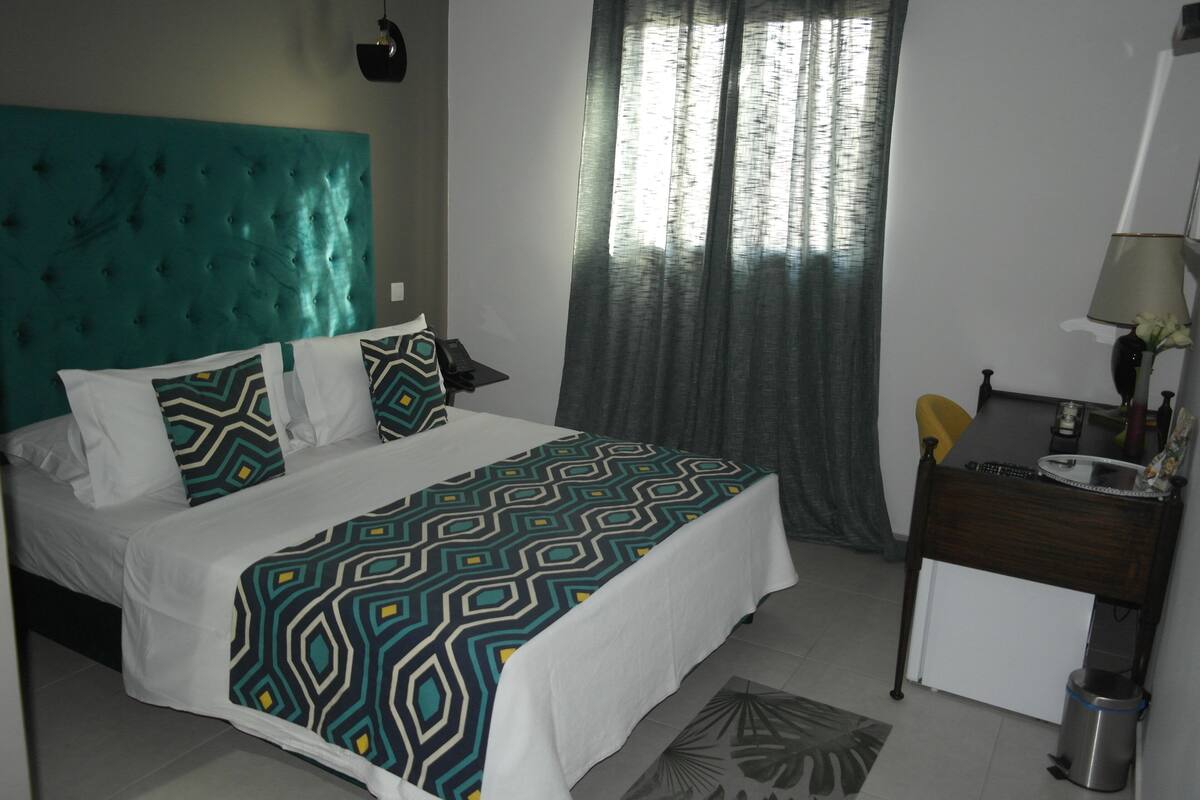 A neatly made bed with a patterned bedspread, a small bedside table with a lamp, and a window with curtains.