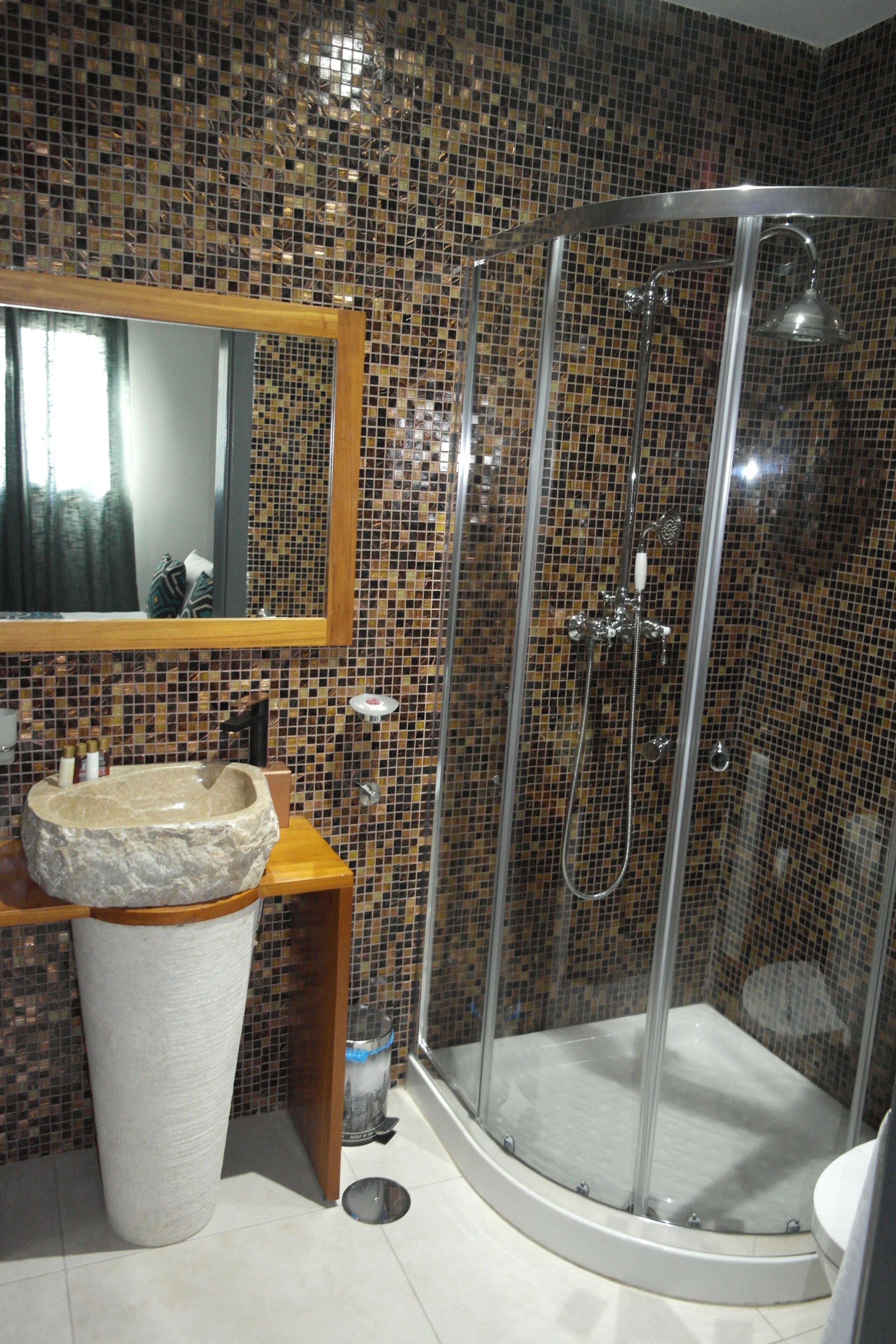 Un baño con pared de mosaicos, tocador de madera con lavamanos y zona de ducha con mampara de cristal.