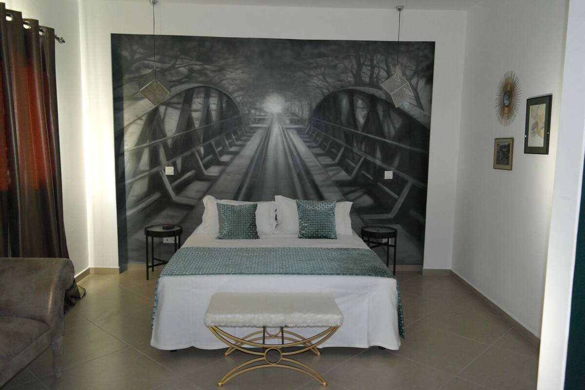 A bedroom with a large bed, a bench, a nightstand, and a wall mural of a bridge.