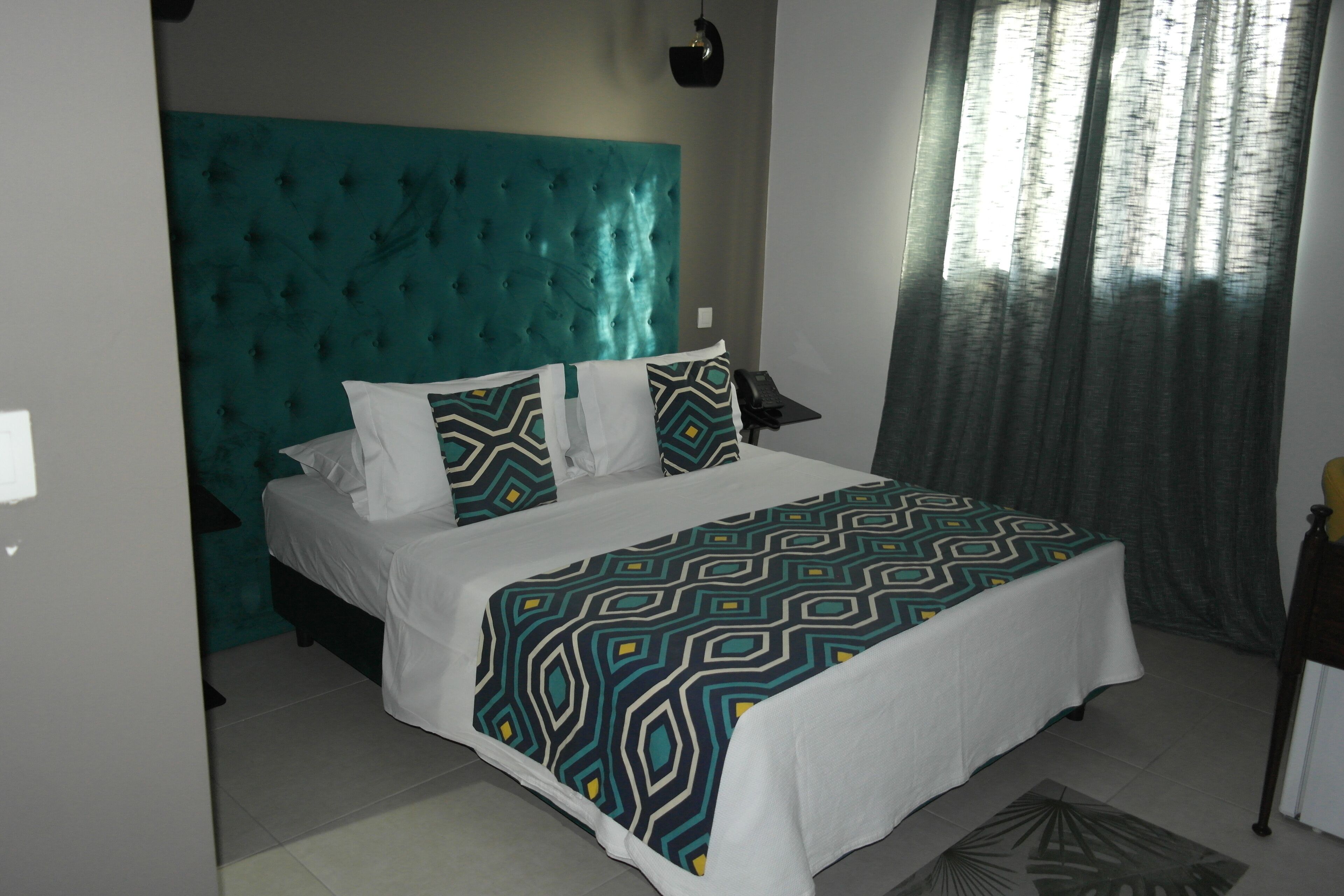 Une chambre à coucher avec une tête de lit capitonnée de couleur turquoise, une couvre-lit à motifs et une vue sur une forêt, visible à travers une fenêtre ornée de rideaux.