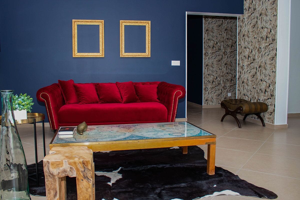 A room with a red sofa, a glass coffee table, a wooden stool, and a black and white rug. There are two framed pictures on the wall and a plant on the left.