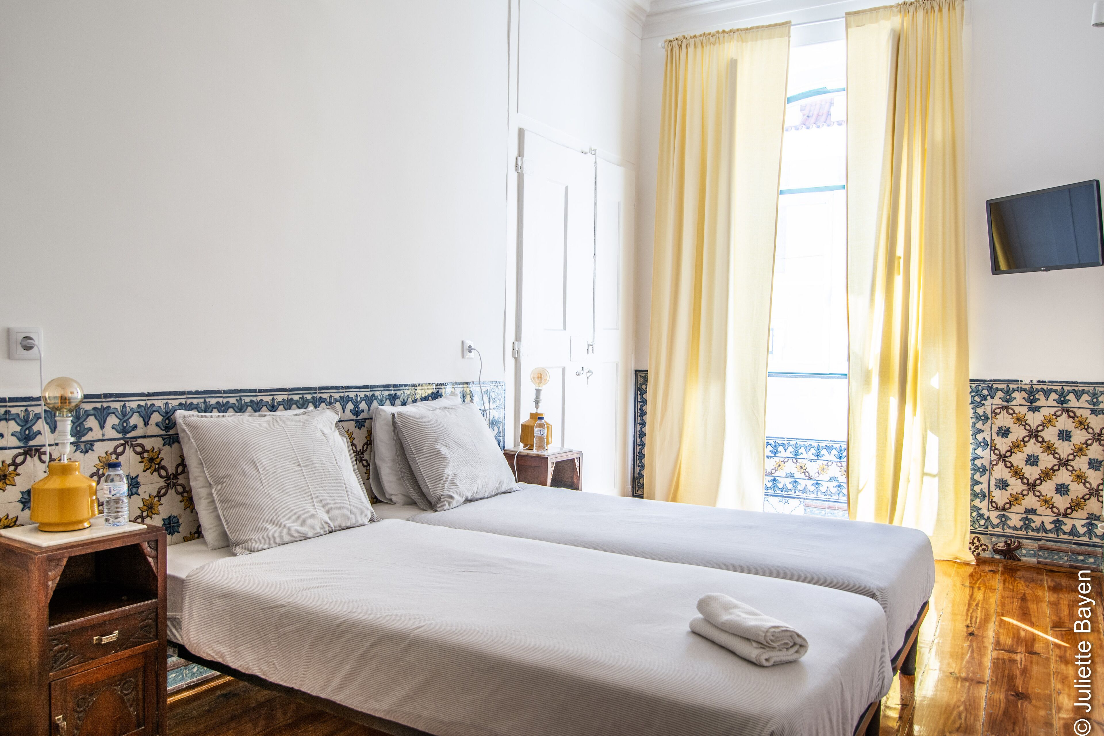 A double bed with white linens, a bedside table with a lamp, a water bottle, and a folded towel.