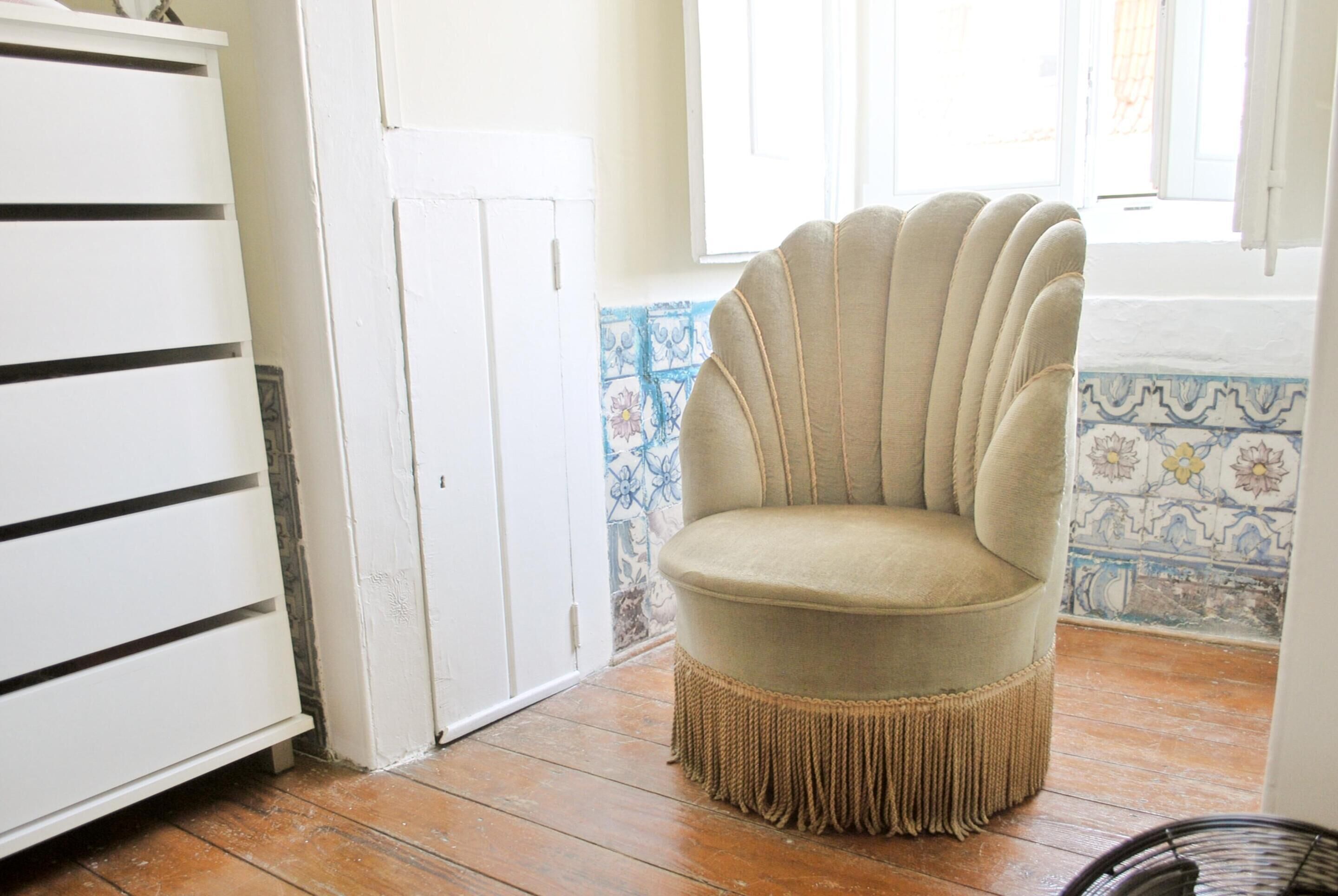 Una habitación con un sillón color beige con flecos, un armario blanco y una pared de azulejos con motivos florales.