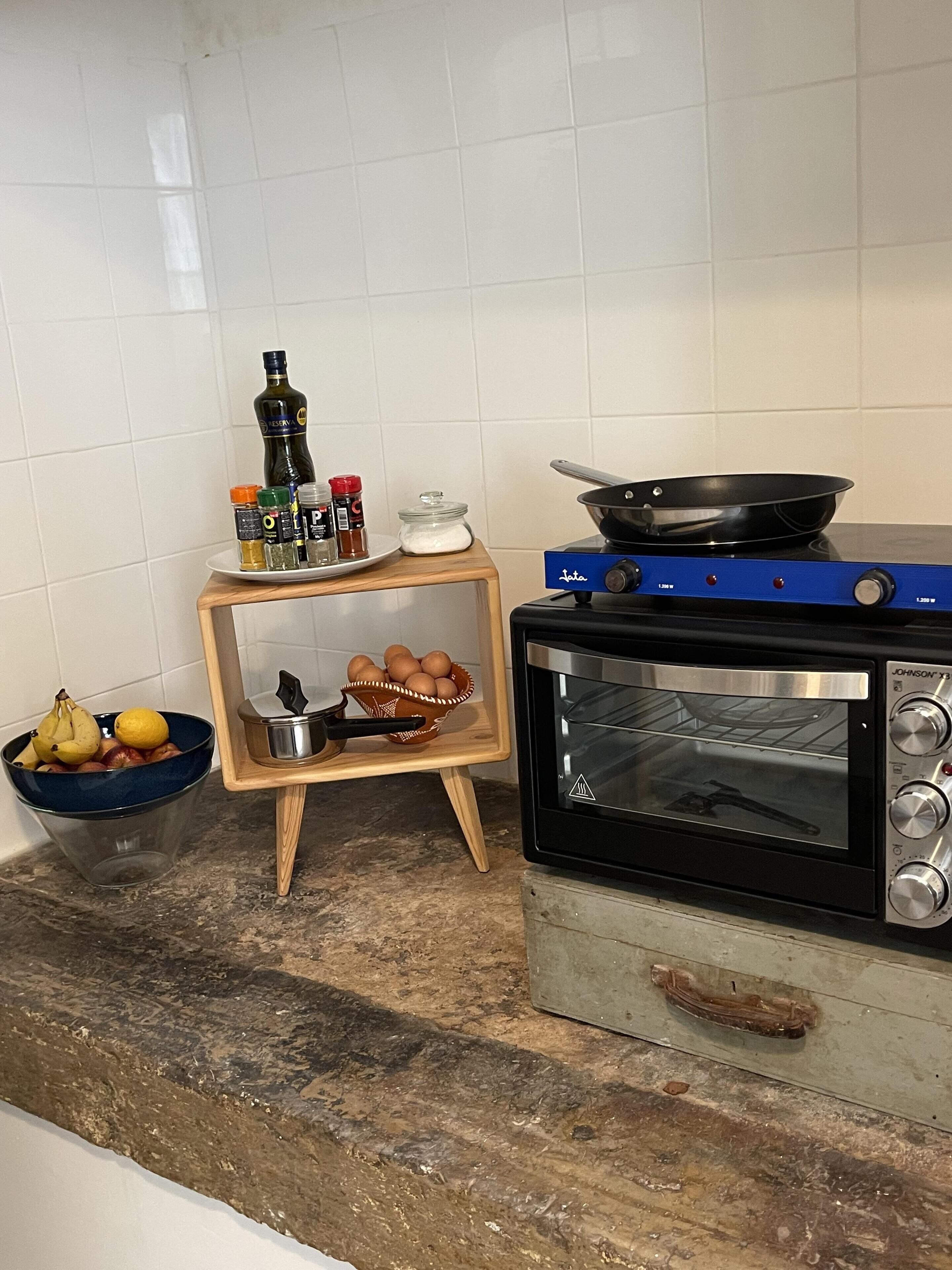 Ett kök med en liten ugn med grillfunktion, ett träställ med kryddor, en skål med frukt och en stekpanna på spisen.