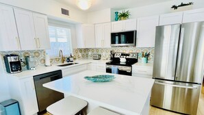 A modern kitchen with white cabinetry, a central island with stools, stainless steel appliances, and a patterned backsplash. - Steps to the Beach, No Car Needed, Completely Permitted Remodel + King Size Bed! (Indian Rocks Beach)