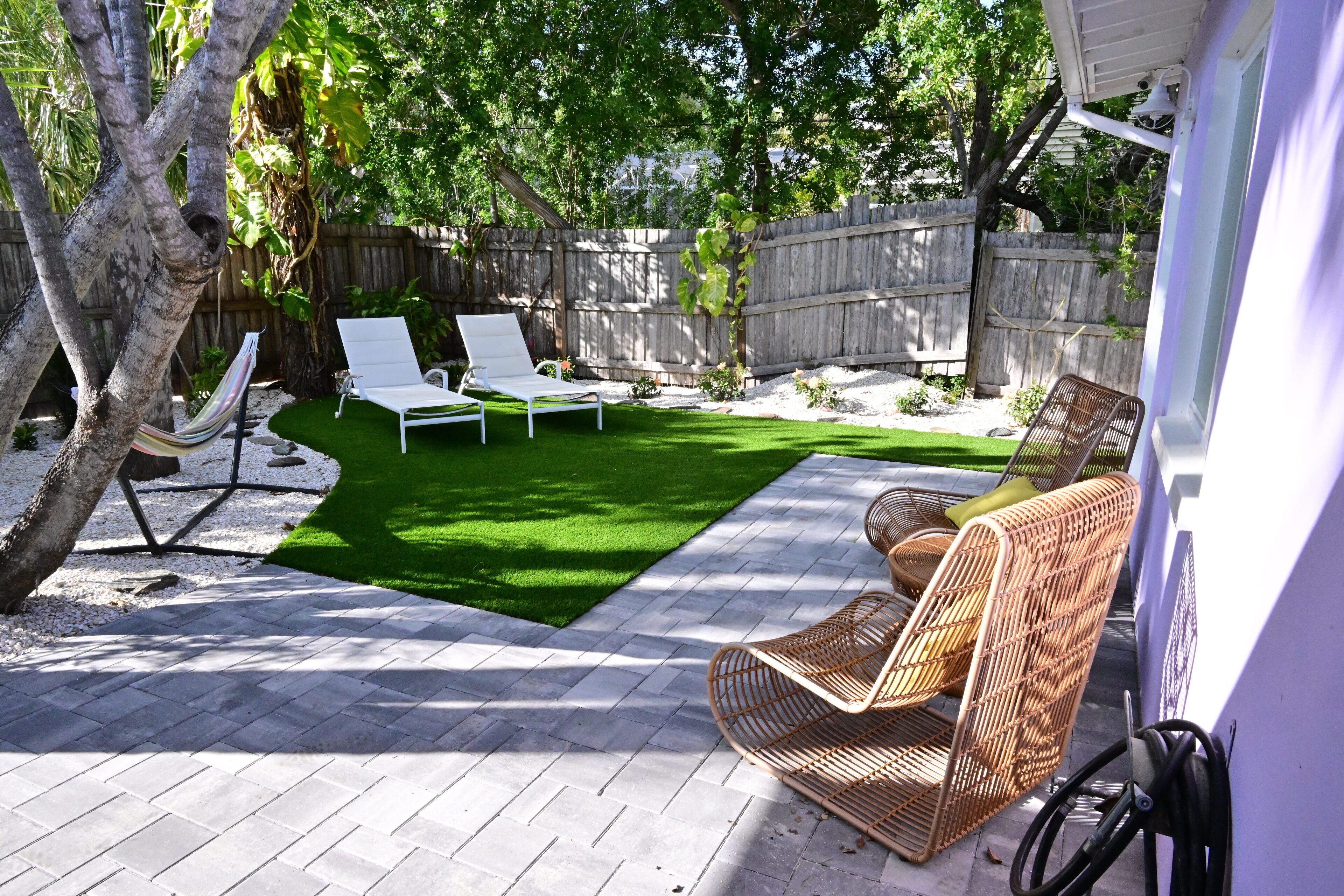 A backyard with a paved patio, two white lounge chairs, a wicker chair, and a hammock.