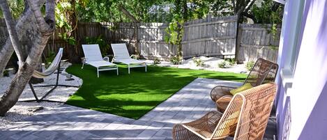 A backyard with a paved patio, two white lounge chairs, a wicker chair, and a hammock.