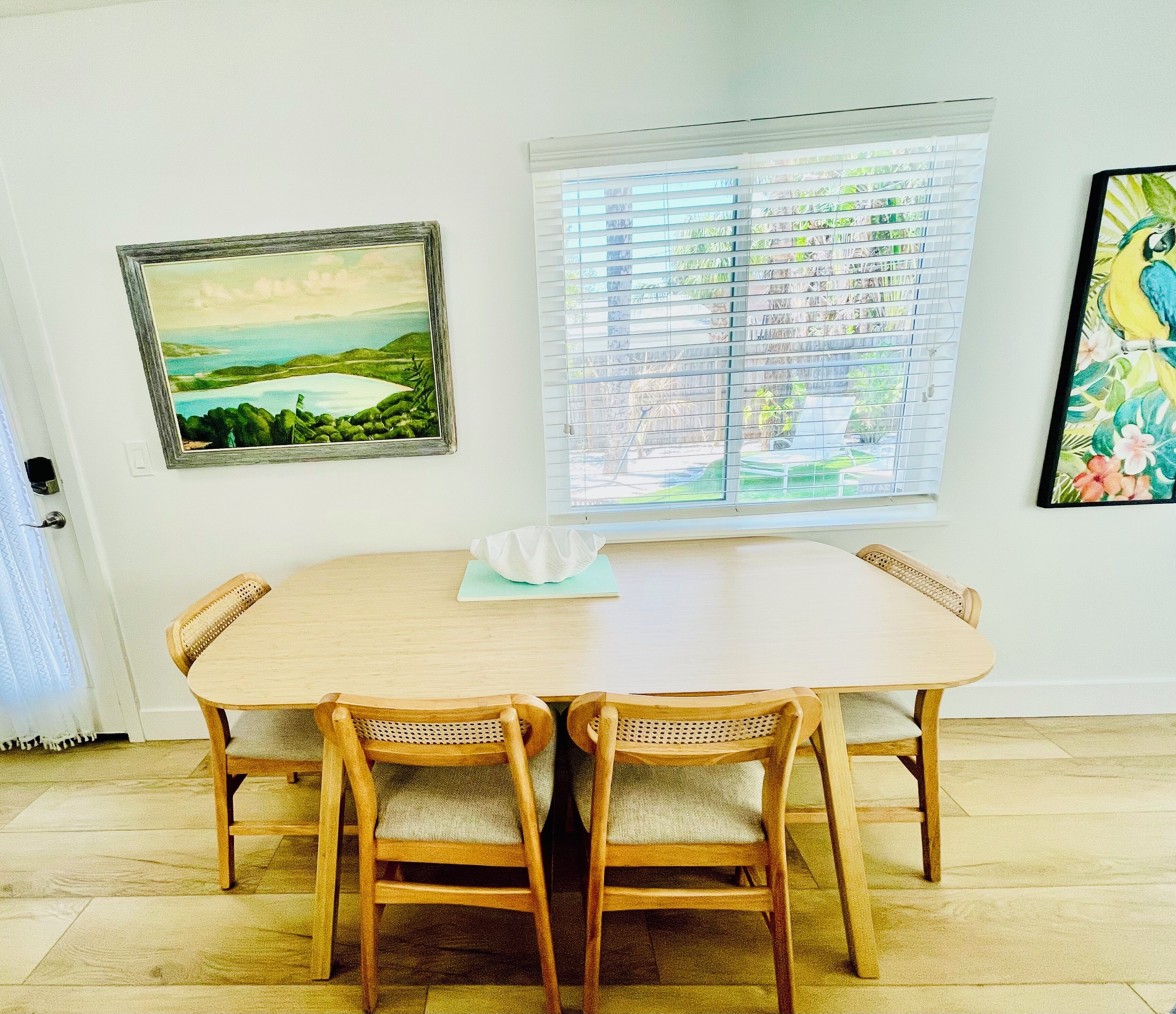 A dining area with a wooden table and chairs, a window with blinds, and two framed artworks on the wall.