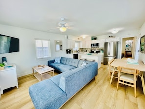 A modern living room with a blue sofa, a dining table with chairs, a kitchen area with white cabinets, and a ceiling fan.