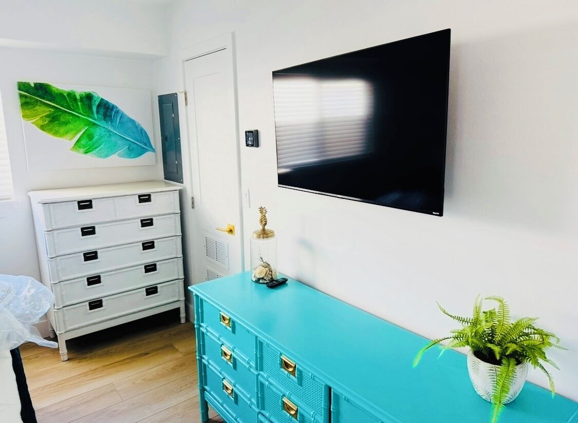 A room with a white dresser, a teal cabinet, a large leaf artwork, a television, and a potted plant.