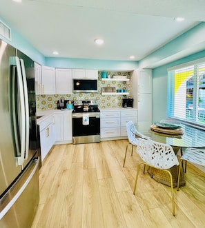 A modern kitchen with white cabinets, a stainless steel refrigerator, a microwave, and a stove. There is a dining table with white chairs and a window with blinds.