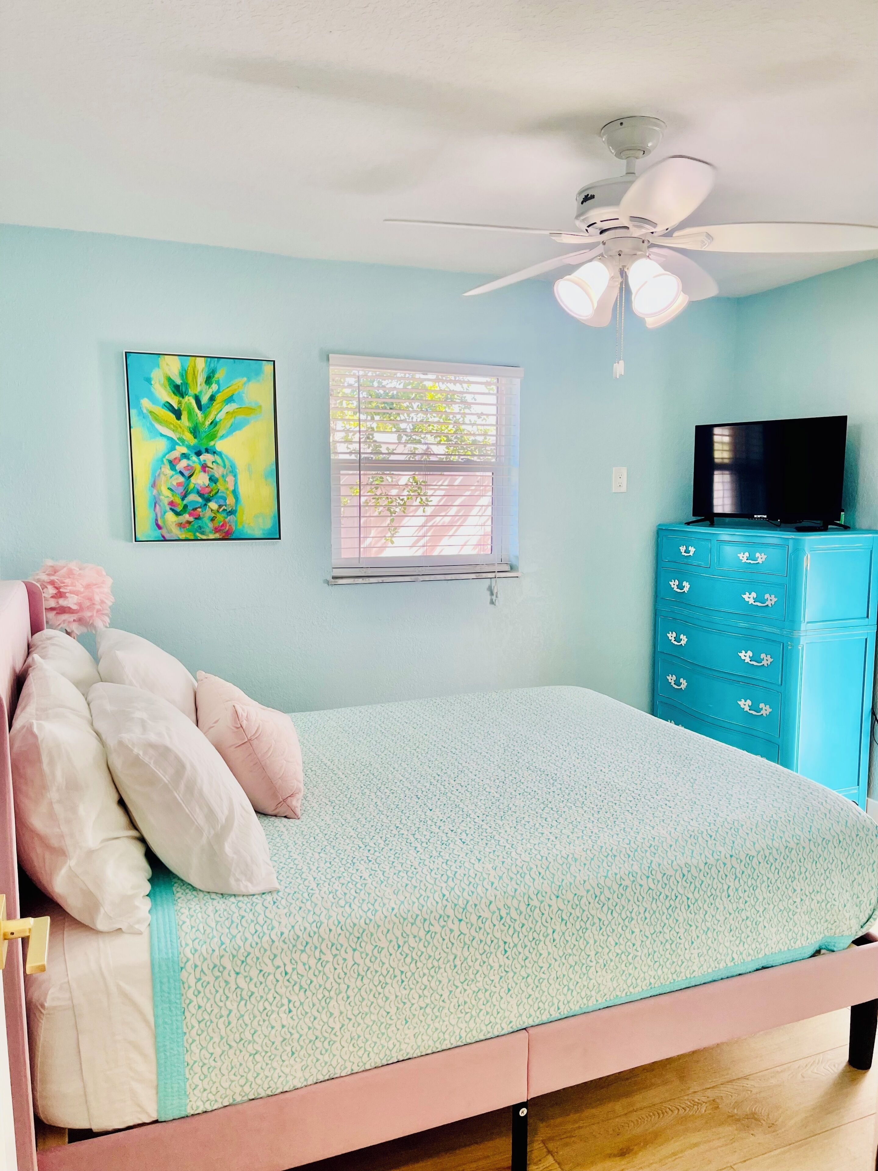 Une chambre à coucher comprenant un lit, un ventilateur de plafond, une télévision, une commode, une fenêtre avec des stores et un tableau.