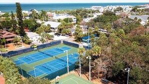 Un court de tennis entouré d’arbres et d’immeubles résidentiels, avec une vue sur l’océan en arrière-plan.