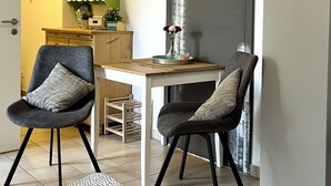 Una sala de estar moderna con dos sillones grises, una mesa de centro de madera y una mesa de comedor con sillas. Se observa una estantería verde con cristalería y un cuadro en la pared.