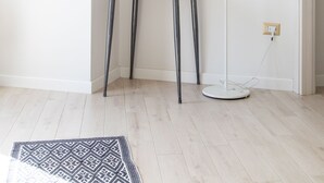 Una habitación con piso blanco, un tapete estampado, una mesa con planta, una lámpara y un espejo en la pared.