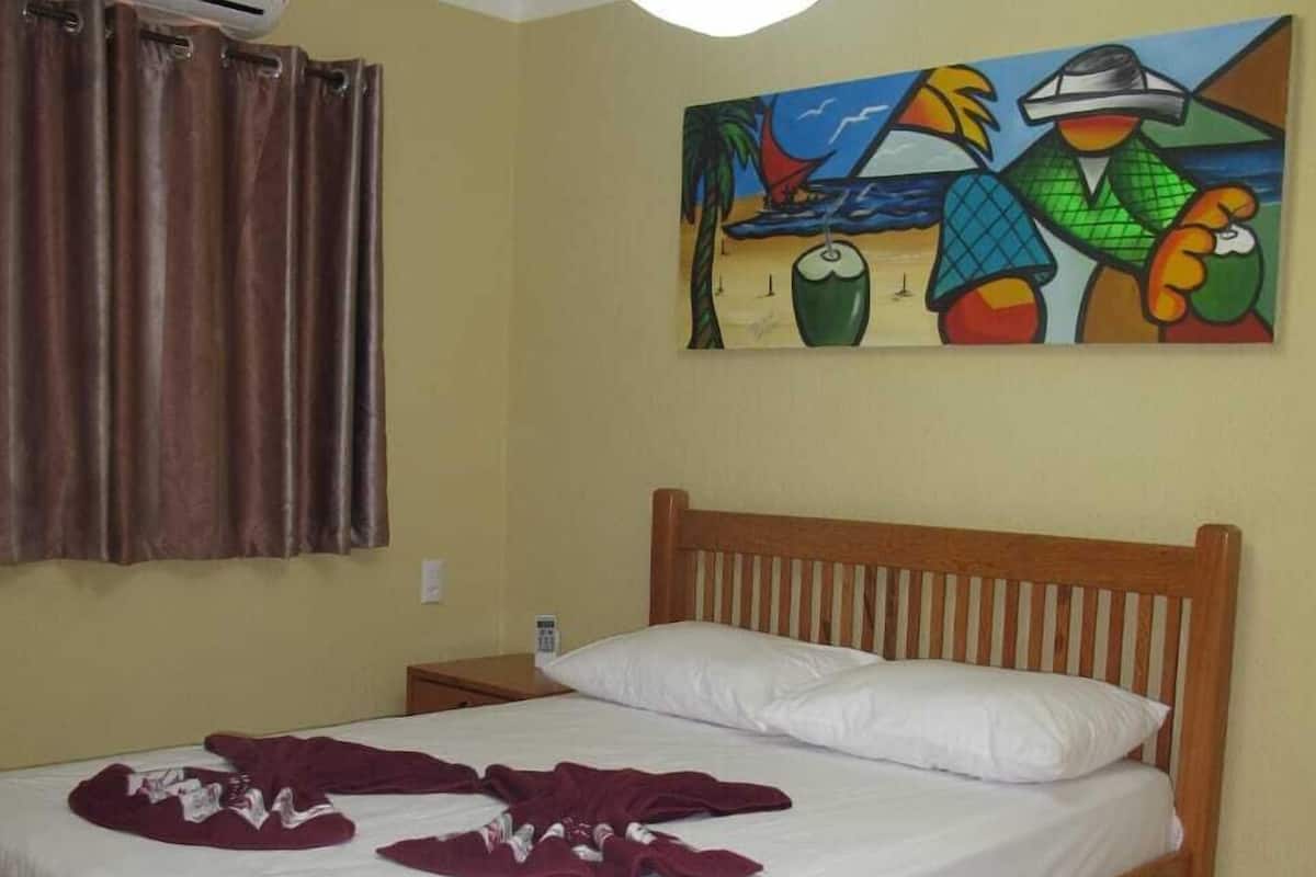 A bedroom with a wooden bed, white bedding, a painting on the wall, and an air conditioning unit.
