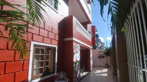 Uma passagem estreita com um edifício de tijolos vermelhos, janelas com molduras brancas e uma pequena área externa com cadeiras e mesa brancas.