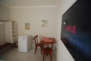 Una habitación pequeña con un refrigerador blanco, una mesa de madera con dos sillas y un televisor de pared que muestra el logotipo de Netflix.