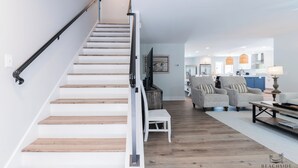 A modern interior with a wooden staircase, a living area with two sofas, a dining table, and a kitchen in the background. - 19 Dove St - Large Pool, Short Walk to Beach! (Hilton Head Island)