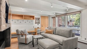 A modern living room with a fireplace, grey sofa, armchair, and ottoman. There is a wooden table with a bowl and a mirror on the wall. - Contemporary Condo in Aspen Core (Aspen)