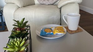 Een witte lederen fauteuil met een kussen met een geometrisch patroon, een witte mok, een schaal met snacks en een vetplant op een witte tafel.