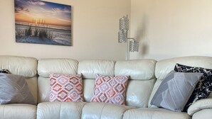 Een woonkamer met een beigekleurige leren bank, gekleurde kussens, een witte salontafel met een plant en een schilderij van een zonsondergang aan de muur.