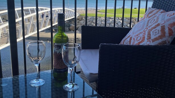 Een balkon met een glazen tafel, twee wijnglazen en een fles wijn. Een rieten stoel met een gestreept kussen staat rechts. Op de achtergrond is een witte brug en een berg te zien.