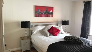 Een slaapkamer met een bed, twee nachtkastjes, een stadslandschap schilderij, een raam met gordijnen en een plafondlamp.