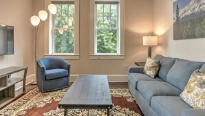 A living room with a sofa, armchair, coffee table, and a TV. There are two windows with a view of trees and a lamp. - Charming, condo #1 in Historic King James Building (Asheville)