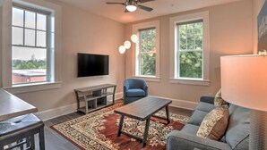 A living room with a flat-screen TV, a ceiling fan, a blue chair, a grey sofa, and a patterned rug. - Charming, condo #1 in Historic King James Building (Asheville)
