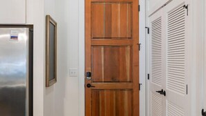 Un couloir avec une porte en bois, des murs blancs, un réfrigérateur en acier inoxydable et une chaise noire.