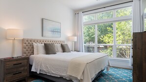 Une chambre avec une grande fenêtre, une tête de lit capitonnée, une table de chevet et un tapis à motifs.