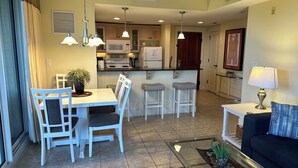 A modern kitchen with white cabinets, a dining area with white chairs, a bar stool, and a living area with a dark sofa and a lamp. - Baytowne Wharf - 4th Floor Corner Apartment (Sandestin)