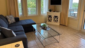 A living room with a sectional sofa, a glass coffee table, a TV on a stand, and large windows with curtains. - Baytowne Wharf - 4th Floor Corner Apartment (Sandestin)
