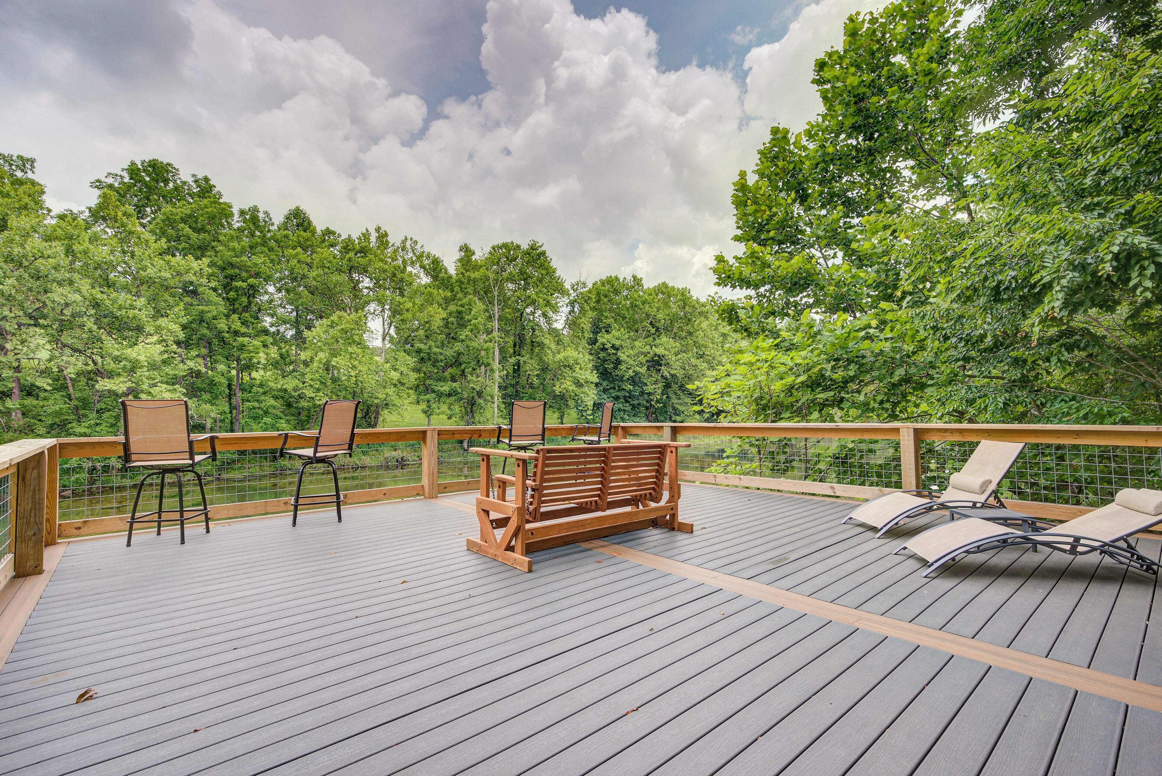 A wooden deck with outdoor furniture, including chairs and a bench, overlooking a forested area.