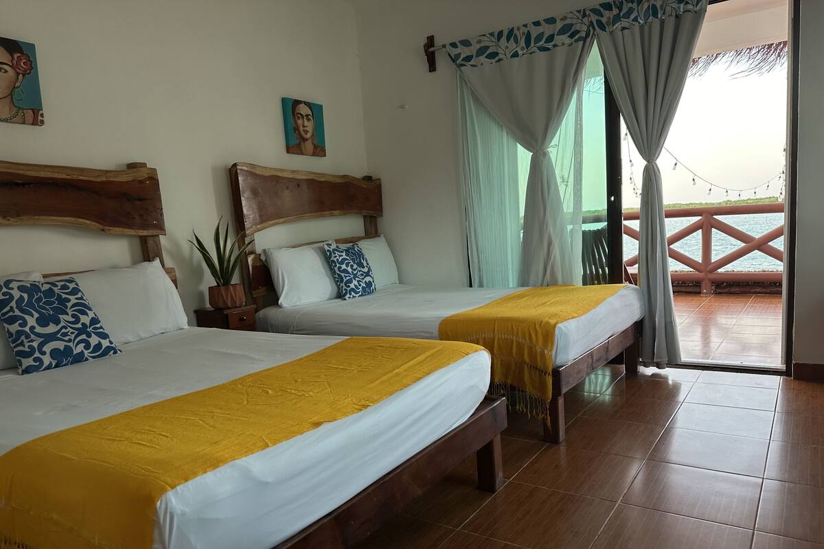 Habitación de hotel con dos camas, cabeceros de madera, ropa de cama blanca y vista al mar desde un balcón.