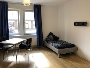 Quarto pequeno e bem iluminado, com uma cama, uma mesa e uma cadeira.