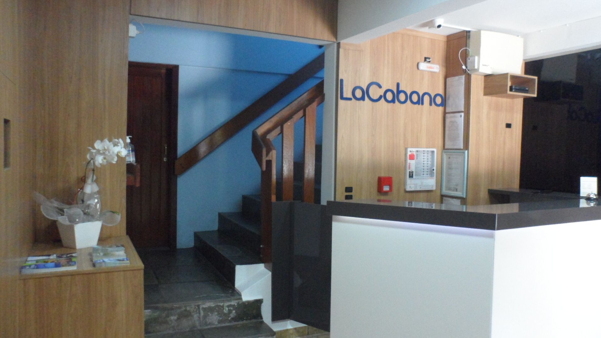 Un área de recepción con una escalera de madera, un letrero de LaCobanah, un mostrador de recepción y un panel de alarma contra incendios.