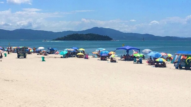 A beach with people, colorful umbrellas, and a distant island. - Apart Hotel La Cabana (Florianópolis)