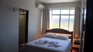 A hotel room with a bed, a window with curtains, an air conditioning unit, and a door. - Apart Hotel La Cabana (Florianópolis)